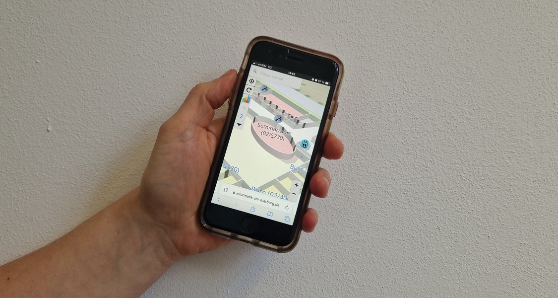 Eine Hand hält ein Smartphone auf dem die Anwendung MINKA einen Seminarraum zeigt