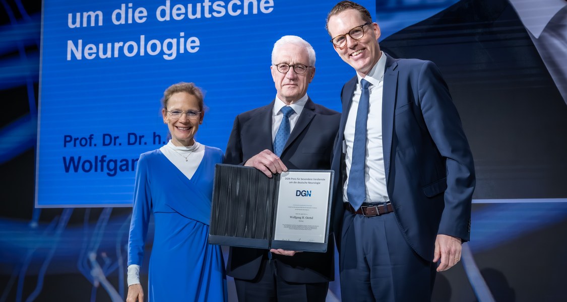 Drei Personen (Frau links in blauem Kleid, mittig Prof. Dr. Wolfgang Oertel, rechts Prof. Dr. Lars Timmermann) mit DGN-Urkunde „besondere Verdienste um die deutsche Neurologie“ auf Bühne in Berlin.