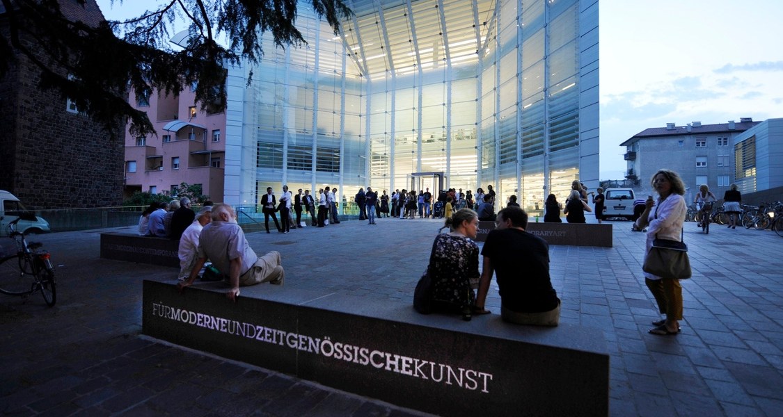 Fassade des Museion in Bozen bei Dämmerung. Zwei Paare sitzend auf einer Bank und eine Gruppe von Menschen stehend vor dem erleuchteten Eingang des Museums.