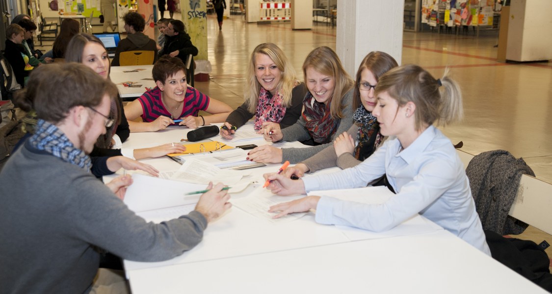 Bild zeigt Studierende, die an einem Tisch im Foyer der Philosophischen Fakultät sitzen und zusammen arbeiten