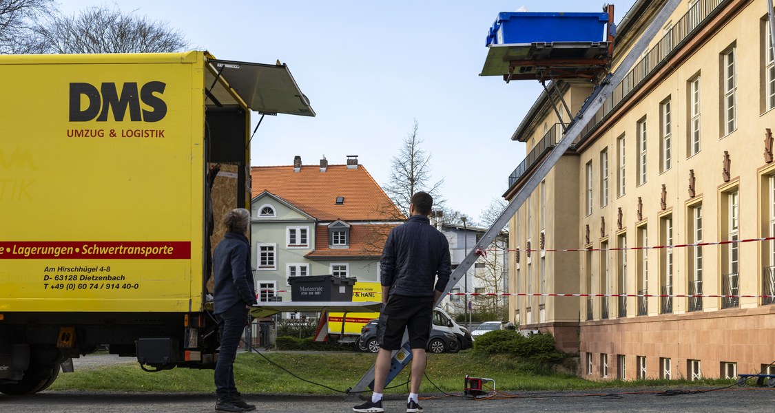 Zwei Personen stehen vor einem Lastenaufzug, der eine blaue Kiste aus dem Dachgeschoss des Kunstgebäudes transportiert. Links ein LKW mit geöffneter Heckklappe.