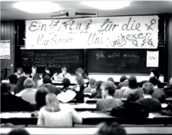 Ein Schwarz-weiß-Bild zeigt Menschen in einem Hörsaal. An der Tafel hängt ein Transparent mit der Aufschrift "Ein ZiFuG für die Marburger Uni. Unihexen".