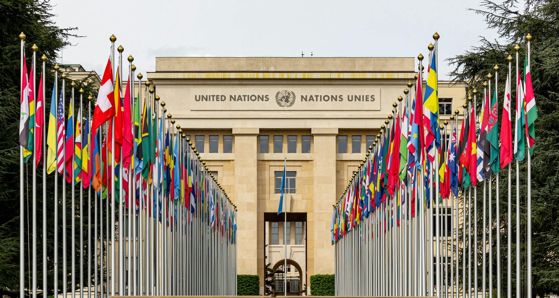 Zu sehen ist das Bürogebäude der Vereinten Nationen in Genf. Vor dem Palais des Nations erstreckt sich die Allée des Nations, mit den Flaggen der UN-Mitgliedstaaten.