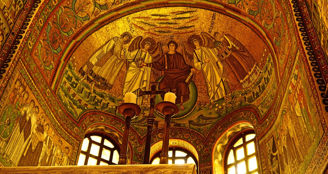 Apsismosaik der Kirche San Vitale in Ravenna, zeigt Jesus Christus als Weltherrscher, neben ihm zwei Engel sowie Ecclesius und Vitalis. Im Vordergrund des Bildes sind das Altarkreuz und zwei Kerzenleuchter zu erkennen. Durch das einfallende Licht leuchten die goldenen Mosaiksteine.