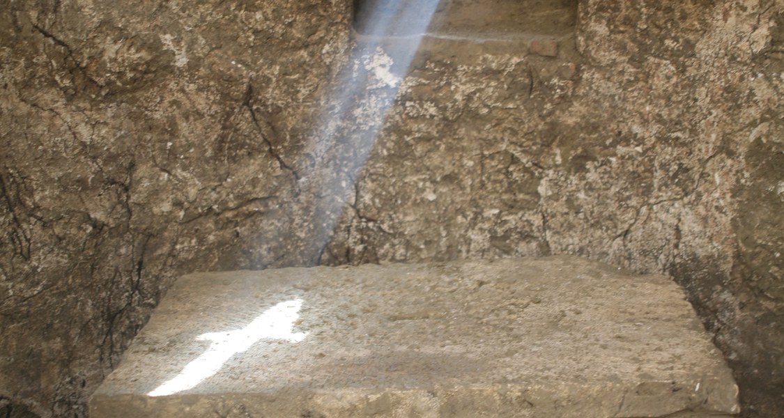 Zu sehen ist die Apsis einer alten Kirche. In der Mauer gibt es eine kleine Auslassung in Kreuzform. Durch das Sonnenlicht, das von außen auf die Kirchenmauer fällt, bildet sich ein Lichtkreuz aus Sonnenlicht auf einem kleinen Altar, der vor der Auslassung in der Mauer steht.