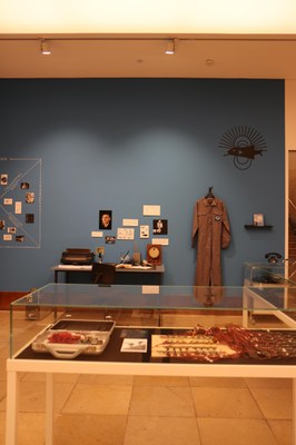 Blick auf ein blaue Wand. Ein Mechaniker Anzug und Bilder an dieser. Davor ein Schreibtisch mit Schreibmaschine und Zetteln. Im Vordergrund eine Glasvitrine mit Kabeln und vermutlich elektronischen Schaltkreisen und Geräten.