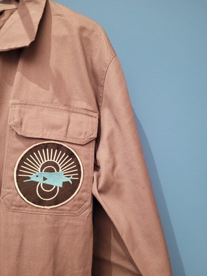 Vor blauer Wand ist ein Ausschnitt von einem Overall zu sehen. Darauf ist ein runder Patch angebracht, der in einfacher Darstellung einen blauen Fisch vor schwarzem Grund zeigt.