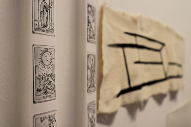 Vor einer weißen Wand, die zur rechten Bildseite hin flüchtet, hängen links zwei schmale Papierstreifen mit schwarzen Drucken. In grober Art ist eine Sonne dargestellt und am unteren Rand steht "Der Mond". In der rechten Bildhälfte ist in einfachen, schwarzen Linien auf Stoff ein liegender Stuhl gemalt.