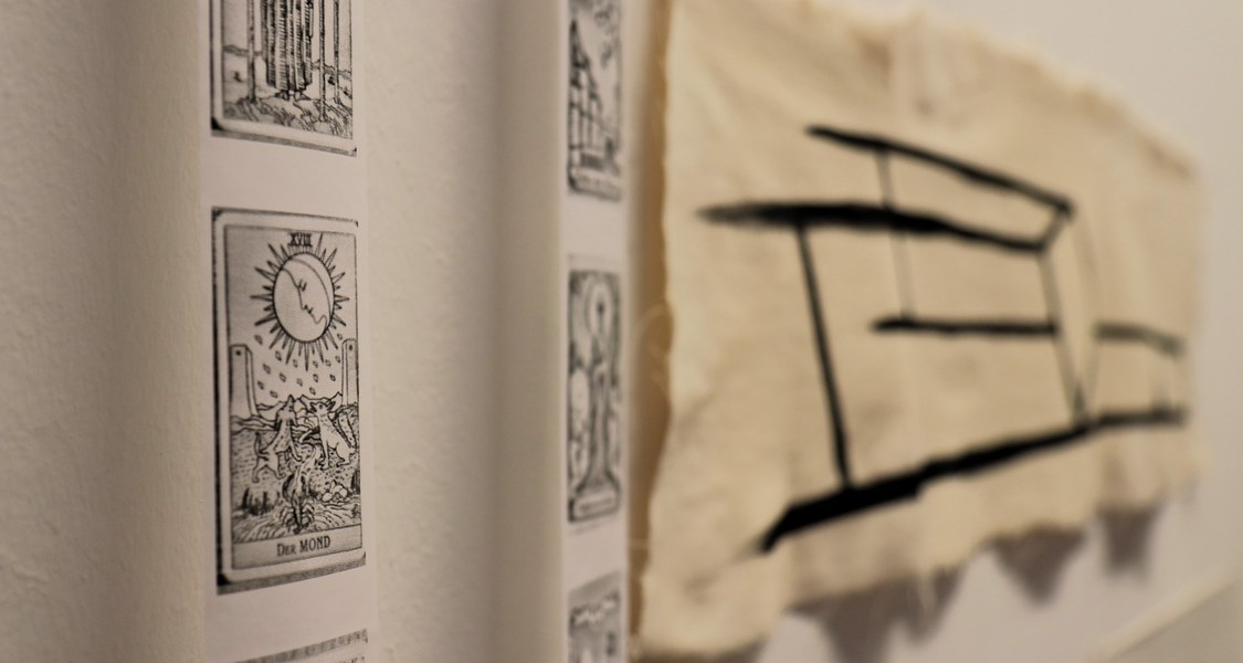 Vor einer weißen Wand, die zur rechten Bildseite hin flüchtet, hängen links zwei schmale Papierstreifen mit schwarzen Drucken. In grober Art ist eine Sonne dargestellt und am unteren Rand steht "Der Mond". In der rechten Bildhälfte ist in einfachen, schwarzen Linien auf Stoff ein liegender Stuhl gemalt.