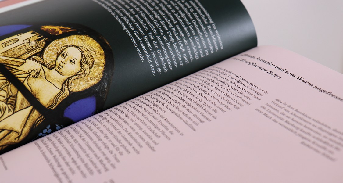 Zwei Buchseiten sind aufgeschlagen. Im Vordergrund ist leicht unscharf Text zu sehen und im Hintergrund ein Ausschnitt eines Glasfensters mit angeschnittener, weiblichen Heiligen.