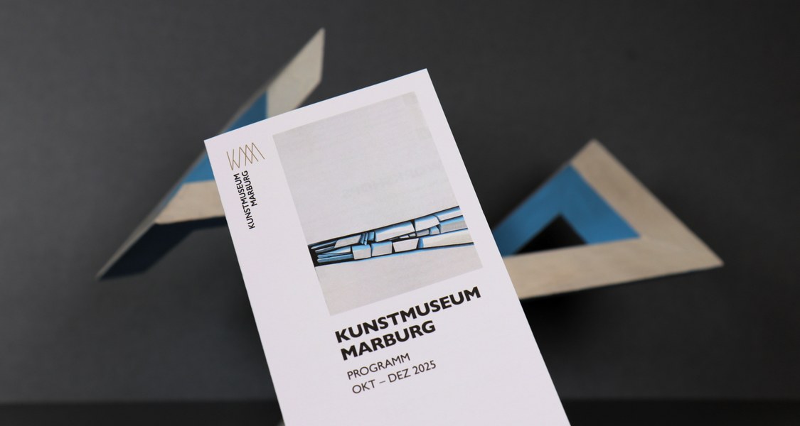 Vorderseite des länglichen Flyers des Quartalsprogramm vom Kunstmuseum vor dunklem Hintergrund. Unscharf im Hintergrund sind die Umrisse eines zckigen, weiß, blauen Gebildes oder Skulptur zu erahnen.