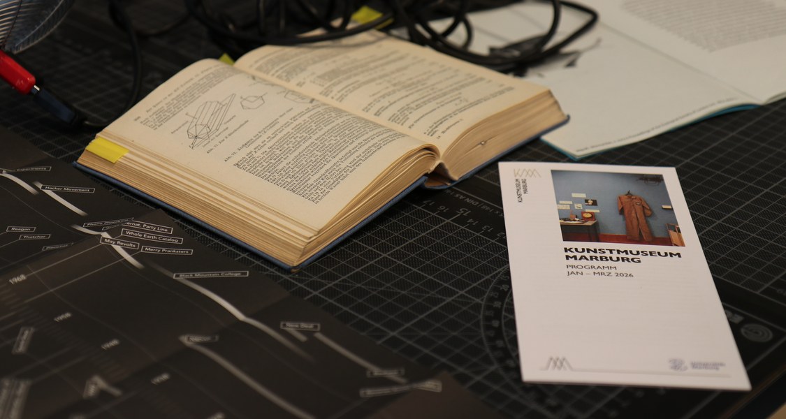 Auf einem Schreibtisch liegt zwischen Büchern und bedruckten Zettel ein Flyer des Kunstmuseums Marburg