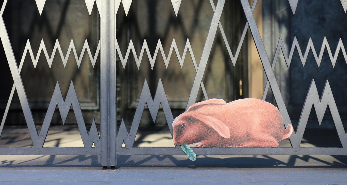 Vor einem schwarzen, gezackten Gittertor sitzt unten rechts ein brauner Hase, der gerade ein Salatblatt frisst.