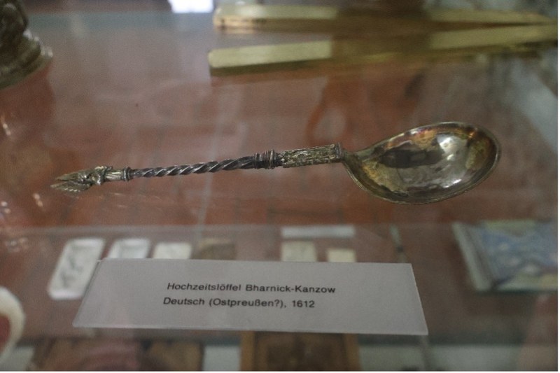 Ein reich verzierter silberner Löffel mit geflochtenen Ornamenten am Stiel liegt in einer Vitrine. Die Beschriftung auf dem Schild lautet: „Hochzeitslöffel, Bharnick-Kanzow, Deutsch (Ostpreußen?), 1612“