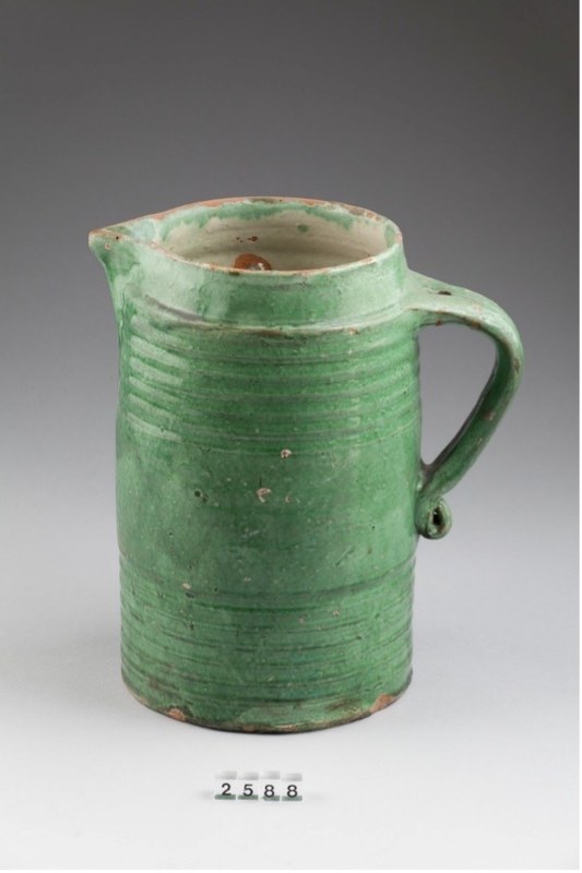 Der hohe zylindrische Keramikkrug hat einen Henkel und eine kleine Ausgusstülle. Man sieht kleine Gebrauchsspuren und helle Stellen; am unteren Rand ist etwas Farbe abgeplatzt.