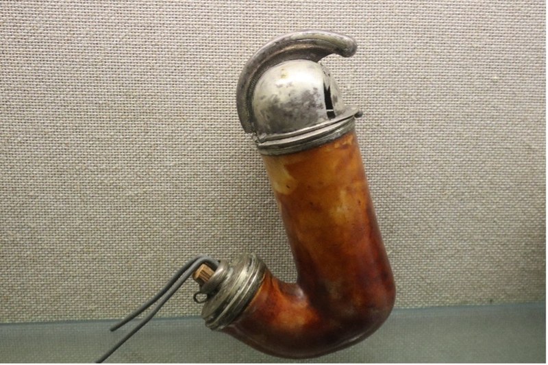 Ein Pfeifenkopf aus braunem, glattem und leicht glänzendem Material in gebogener Form wie ein Haken oder ein „J“ hat oben eine silberne Metallkappe mit gebogenem Metallbügel. Seitlich unten ist eine kleine Öffnung, mit einem kurzen Ansatz und einem Metallring, an dem  zwei Metallstäbe zum Aufstellen des Pfeifenkopfs befestigt sind.