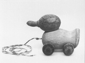 Holzente mit rundem Kopf, einem Schnabel wie ein kleiner Knauf, rundlichem Körper und Rädern; vor der Ente liegt eine Schnur, mit der sie gezogen werden kann.