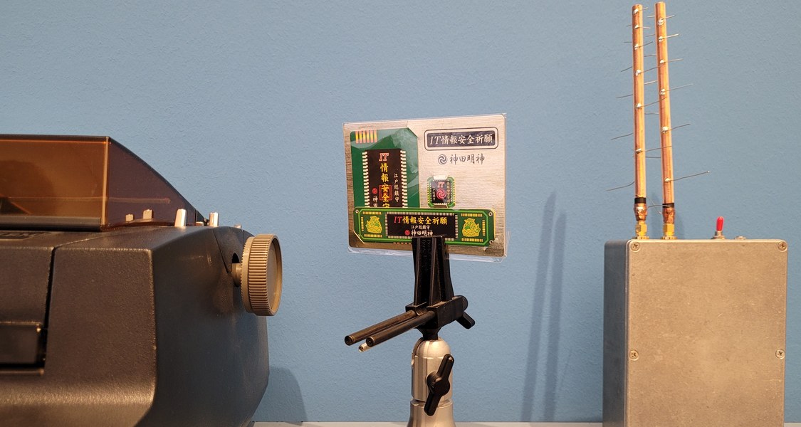 Auf einem Tisch vor blauer Wand steht links eine Schreibmaschine, in der Mitte ein glockenförmiger Kartenhalter, indem eine Visitenkarte mit asiatischen Schriftzeichen steckt und ganz rechts ein rechteckiger grauer Kasten aus dem zwei goldgelbe Antennen emporragen.