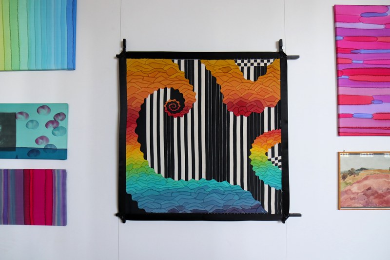An einer Wand hängt zentral ein buntes, quadratisches Bild aus Stoff. Rechts und links am Rand kleine, graphische Bilder.