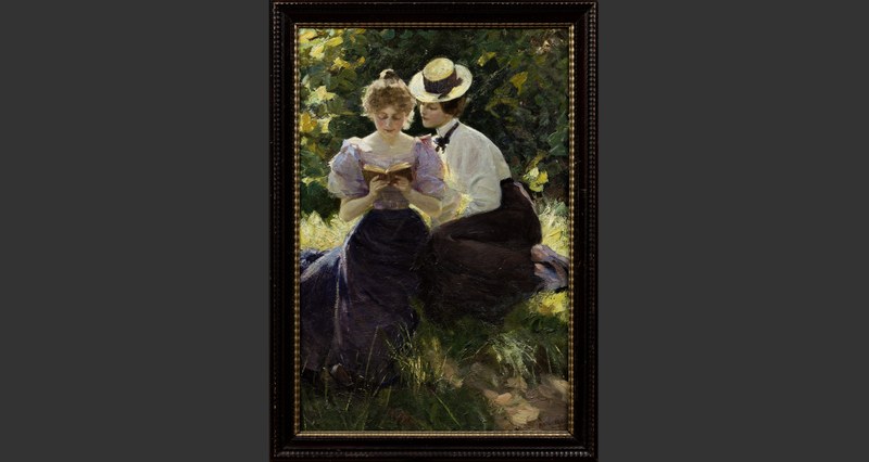 Zwei Frauen sitzen aneinandergeschmiegt im Grünen und lesen aus einem Buch.