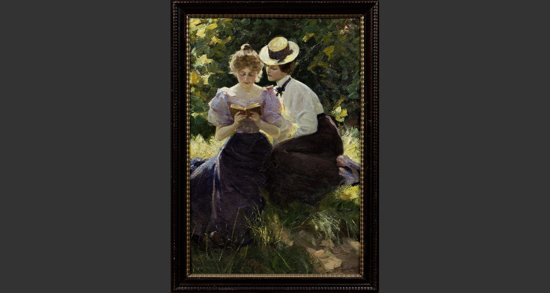 Zwei Frauen sitzen aneinandergeschmiegt im Grünen und lesen aus einem Buch.