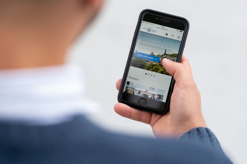 Eine Hand, die ein Smartphone in der Hand hält. Auf dem Display ist die Webseite der Universität Marburg zu sehen.