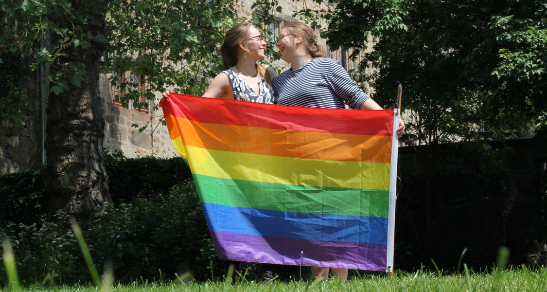 Zwei Frauen halten an einem sonnigen Tag eine Regenbogenflagge in den Händen und schauen sich verliebt an.