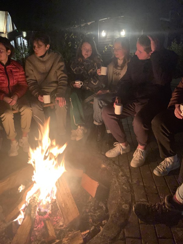 Die Mitbewohner*innen sitzen in warmen Jacken und mit weißen Glühweintassen vor einem Lagerfeuer mit lodernden Flammen.