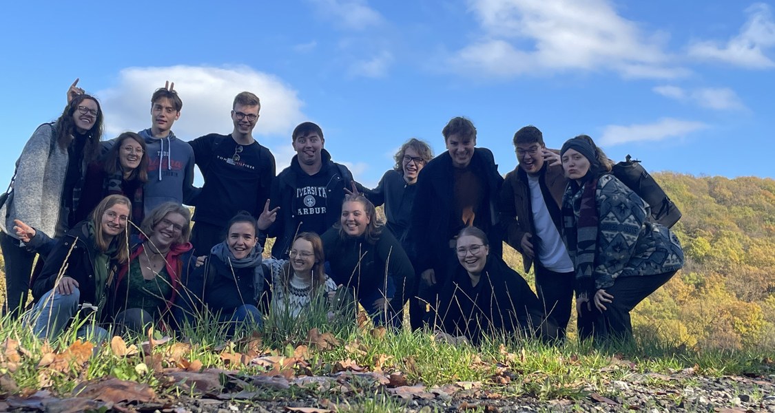 Die Mitbewohner*innen sind vor einer sonnigen Hügellandschaft zum Gruppenphoto aufgestellt.