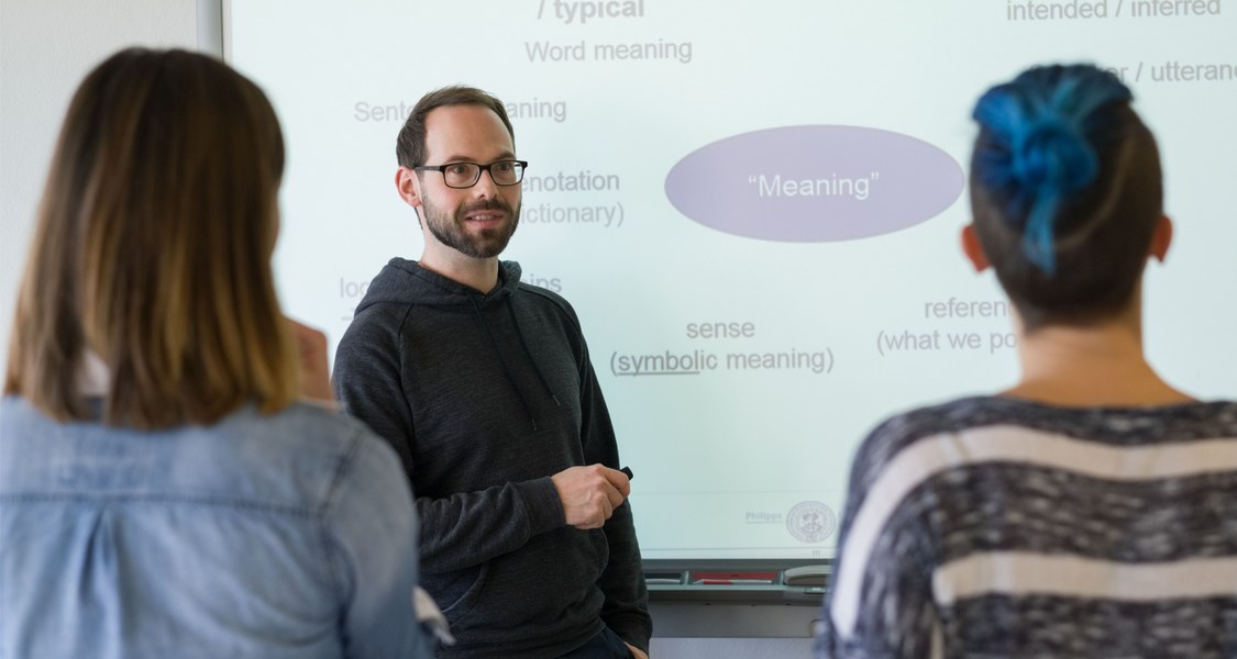 Drei Studierende stehen vor einem Whiteboard, auf welches eine PowerPoint-Folie projiziert wird. Darauf sind verschiedene englische Wörter zu lesen. Der eine Student trägt Brille und Bart, hält einen Pointer in der Hand und scheint den anderen beiden, die mit dem Rücken zur Kamera stehen, etwas zu erklären.