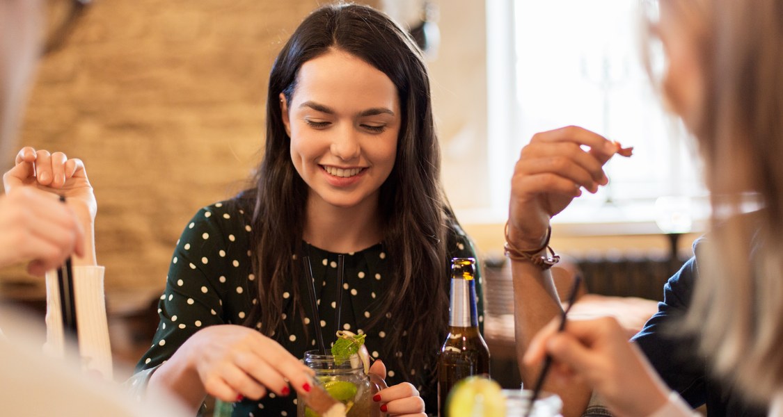 Eine junge Frau sitzt lächelnd mit mehreren Leuten in einer Gaststätte und hat Häppchen, Limonade und eine Flasche Bier vor sich.