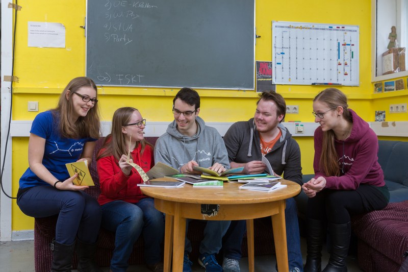 Fünf Studierende sitzen in einem Fachschaftsraum und blicken gemeinsam lachend in verschiedene Broschüren.