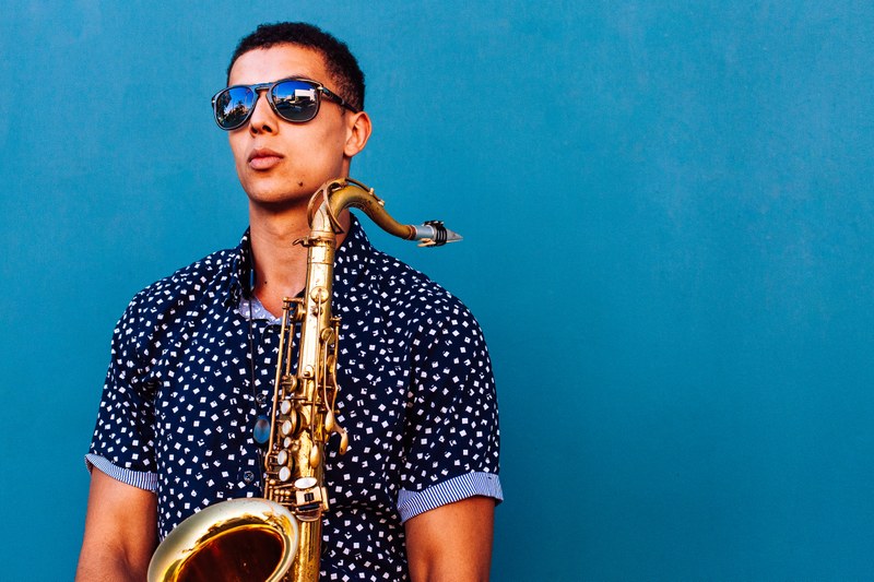Ein Mann mit Sonnenbrille und Saxophon vor blauem Hintergrund.