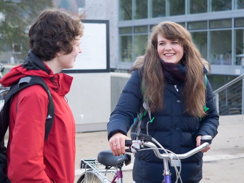 Zwei Studierende stehen in Winterkleidung vor der Mensa Erlenring. Die rechte Person hält ihr Fahrrad am Lenker vor sich.