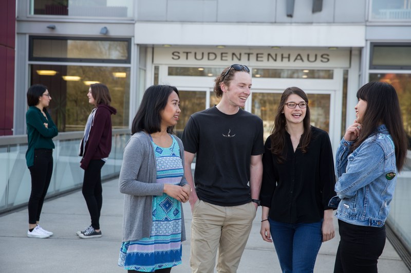 Vier Studierende stehen lachend auf der Mensa-Brücke vor dem Eingang zum Studentenhaus.
