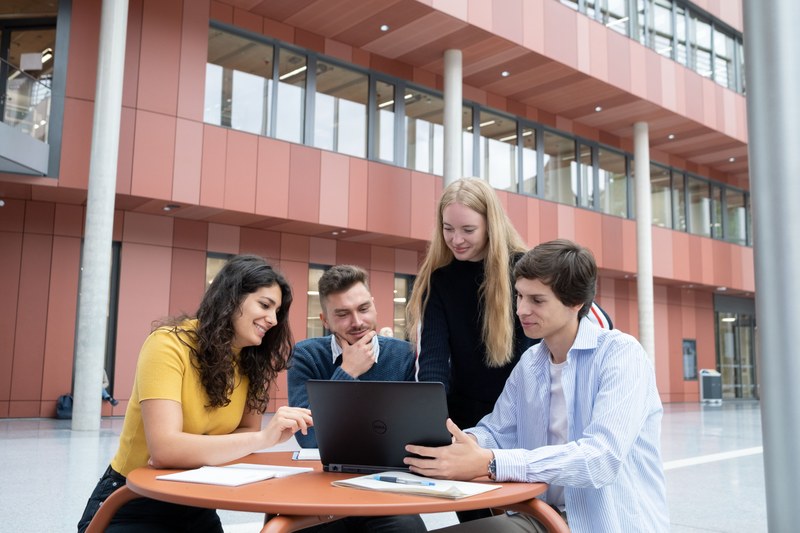 Vier Studierende schauen gemeinsam auf den Bildschirm eines Laptops. Sie sitzen im Atrium der Universitätsbibliothek.
