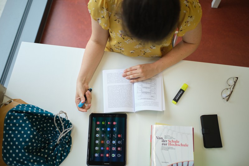 Eine Studentin arbeitet am Schreibtisch in einer Einzelkabine der Universitätsbibliothek. Vor ihr liegen Tablet, Stiftmarker, Handy, Brille, ein Notizbuch und ein Buch aus dem Bestand der UB.