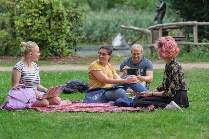 Vier Studierende sitzen einer Picknickdecke auf einer Wiese im Alten Botanischen Garten und besprechen ihre Lernunterlagen.