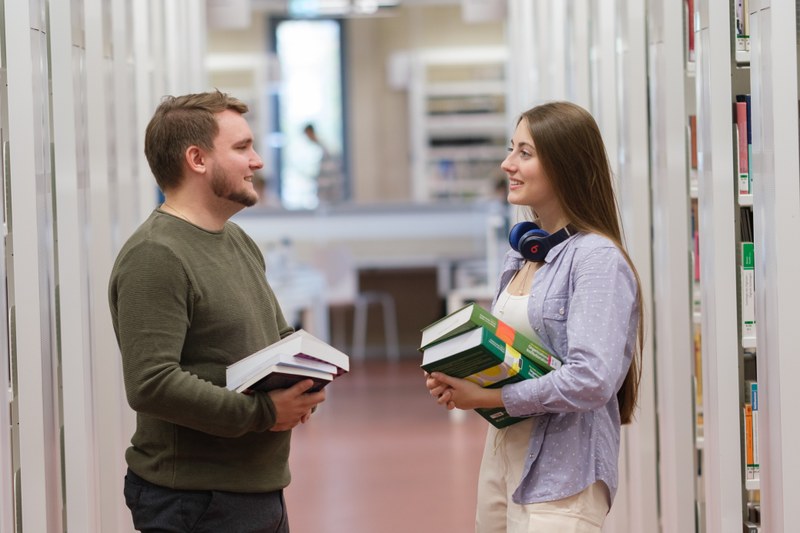 Zwei Studierende stehen mit schweren Büchern auf dem Arm zwischen zwei Regalreihen in der Universitätsbibliothek und unterhalten sich.