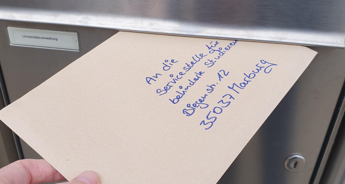 Das Bild zeigt den Briefkasten der Universitätsverwaltung, in diesen gerade ein Brief mit der Adresse der SBS gesteckt wird.