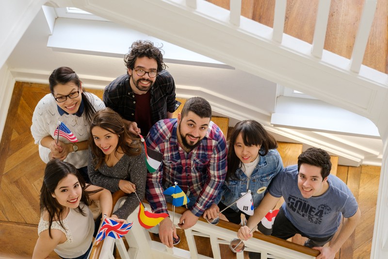 Eine Gruppe Studierender steht mit bunten Länderflaggen in den Händen lachend im Treppenhaus.