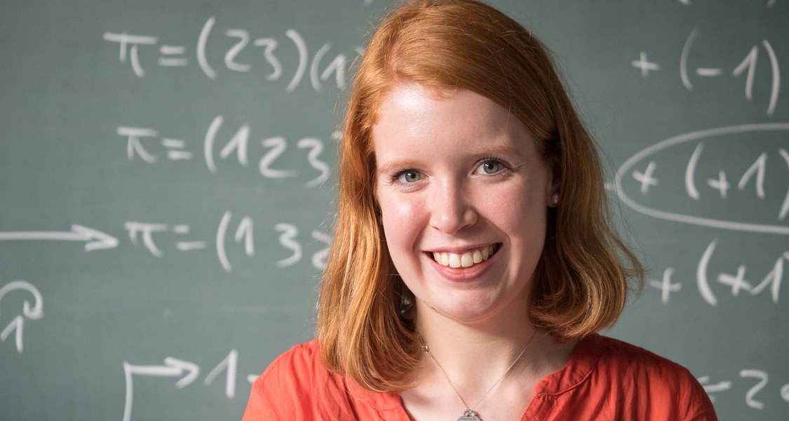 Eine Studentin steht lächelnd vor einer grünen Tafel, die mit mathematischen Formeln beschrieben ist.