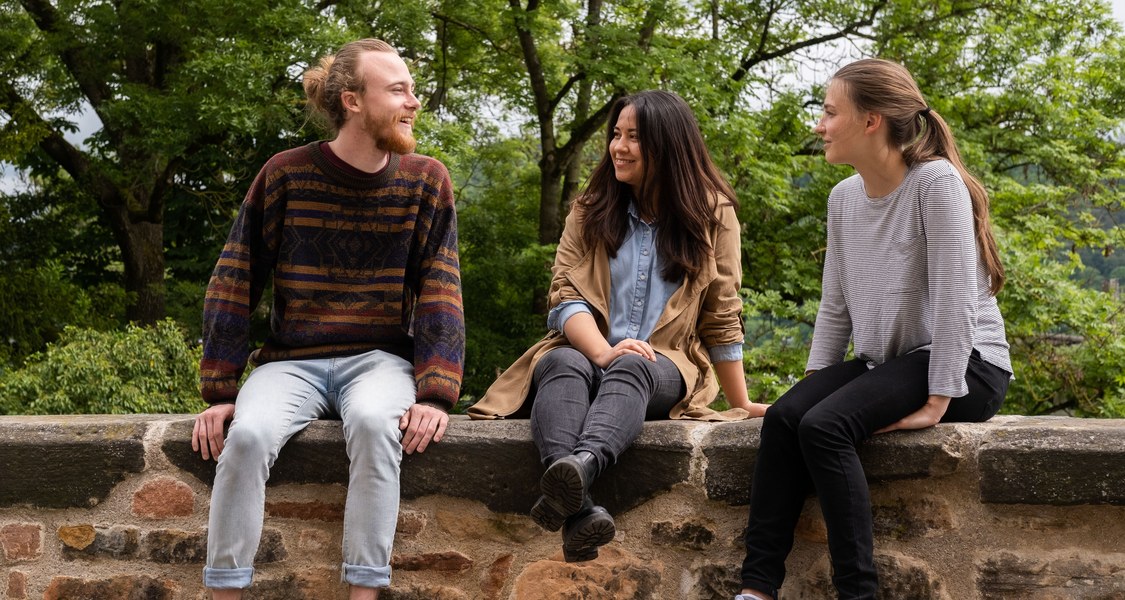 Drei Studierende unterhalten sich auf einer Mauer sitzend.