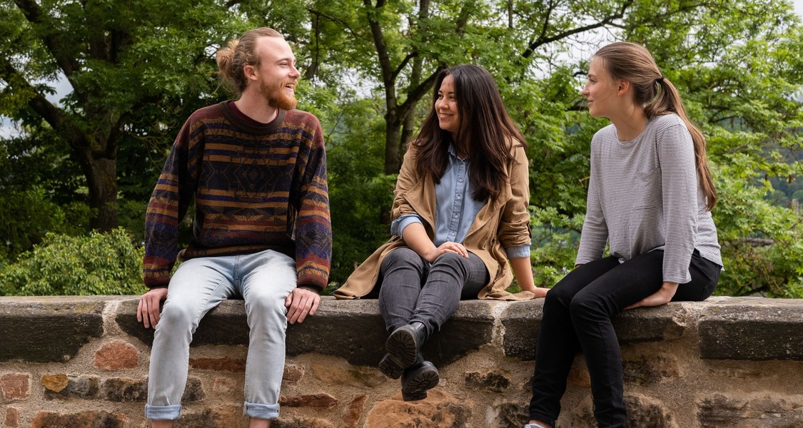 Drei Studierende unterhalten sich auf einer Mauer sitzend.