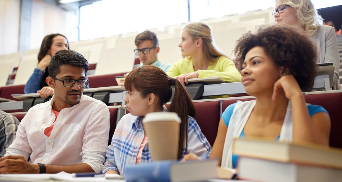 Studierende verschiedenen Ethnien sitzen in einem Hörsaal. Vor ihnen auf den Tischen liegen Bücher und Schreibunterlagen. Sie unterhalten sich miteinander.