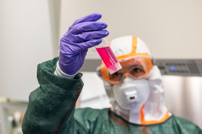 Eine Person im Schutzanzug hält ein Reagenzglas mit pinker Flüssigkeit in die Höhe.