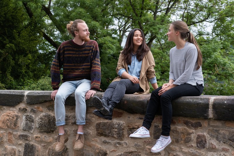 Drei Studierende sitzen auf einer steinernen Mauer und lachen miteinander.