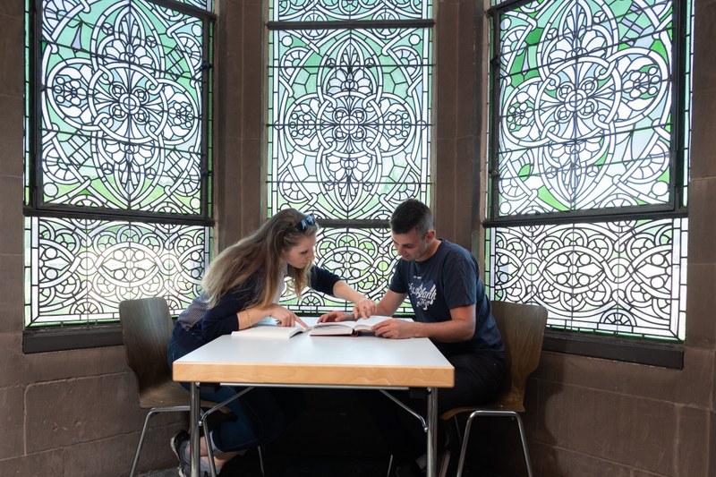 Zwei Studierende lernen über Bücher gebeugt zusammen in einer Glasfensternische der Alten Universität.