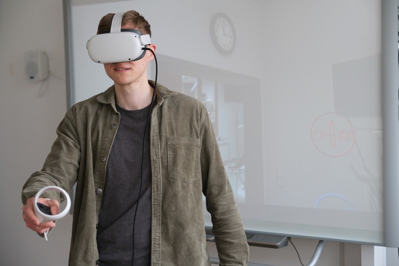 Ein Student hat eine VR-Brille auf dem Kopf und einen Joystick in der Hand.