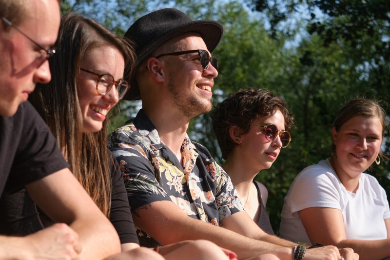 Fünf Studierende sitzen barfuß an der Lahn und unterhalten sich lächelnd.